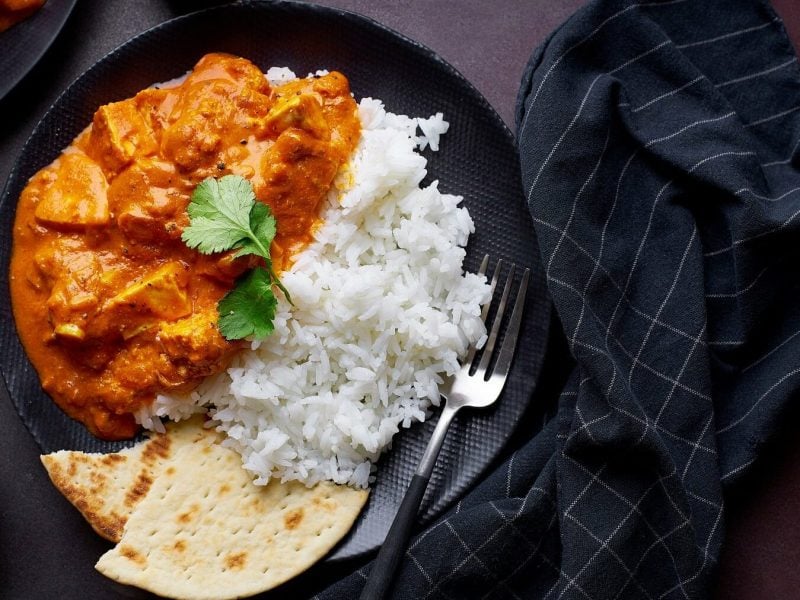 Tofu Tikka Masala mit Reis und Brot auf einem schwarzen Teller mit einer Gabel auf einem dunklen Tisch neben einem schwarzen Tuch mit streifen.