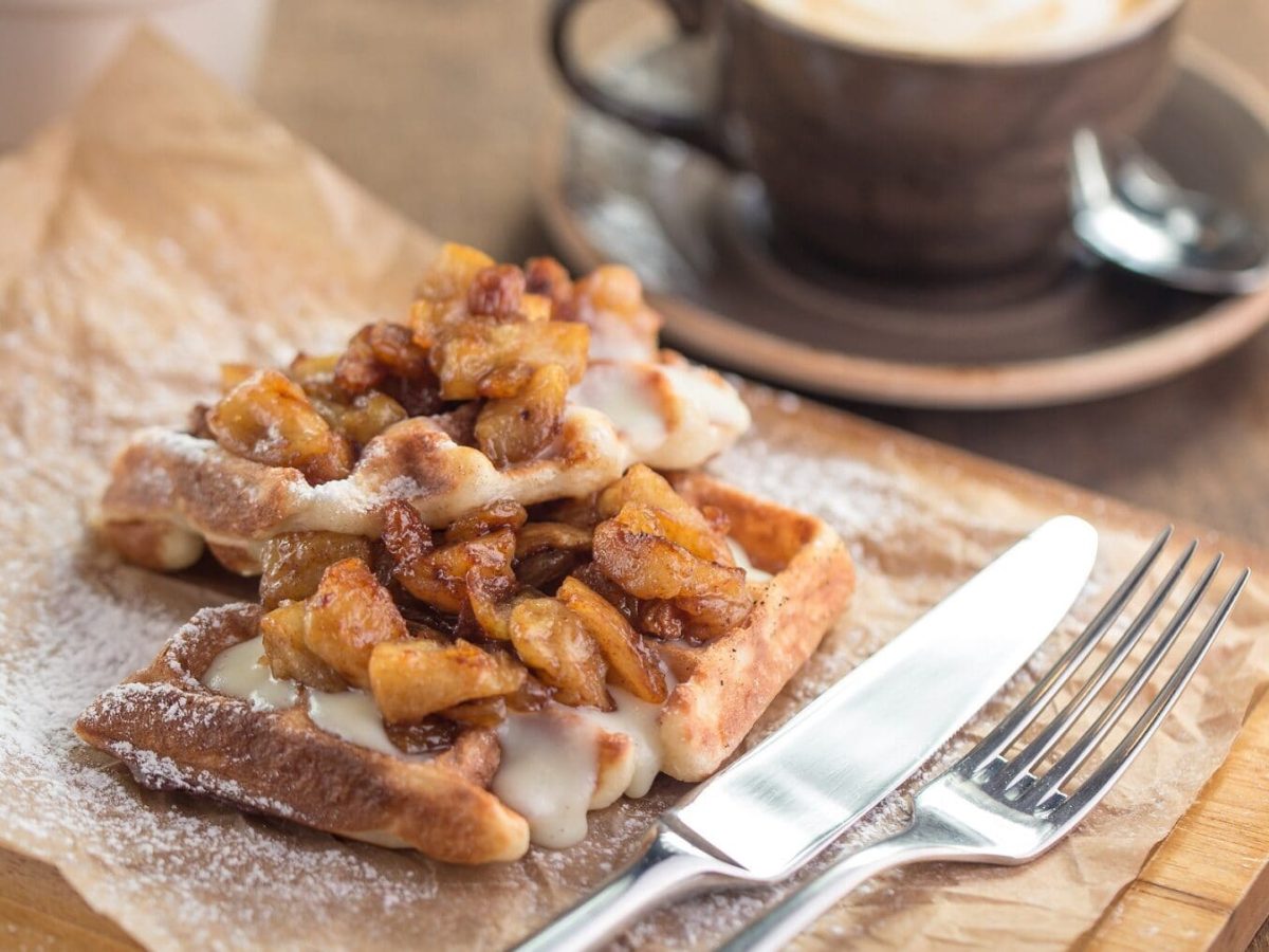 Zwei Vanille-Waffeln mit Bratapfel liegen auf einem braunen Papier auf einem Holzbrett. Neben den Waffeln liegen Messer und Gabel. Im Hintergrund steht ein weißer Becher und eine braune Tasse mit Kaffee. Die Tasse steht auf einer passenden, braunen Untertasse.