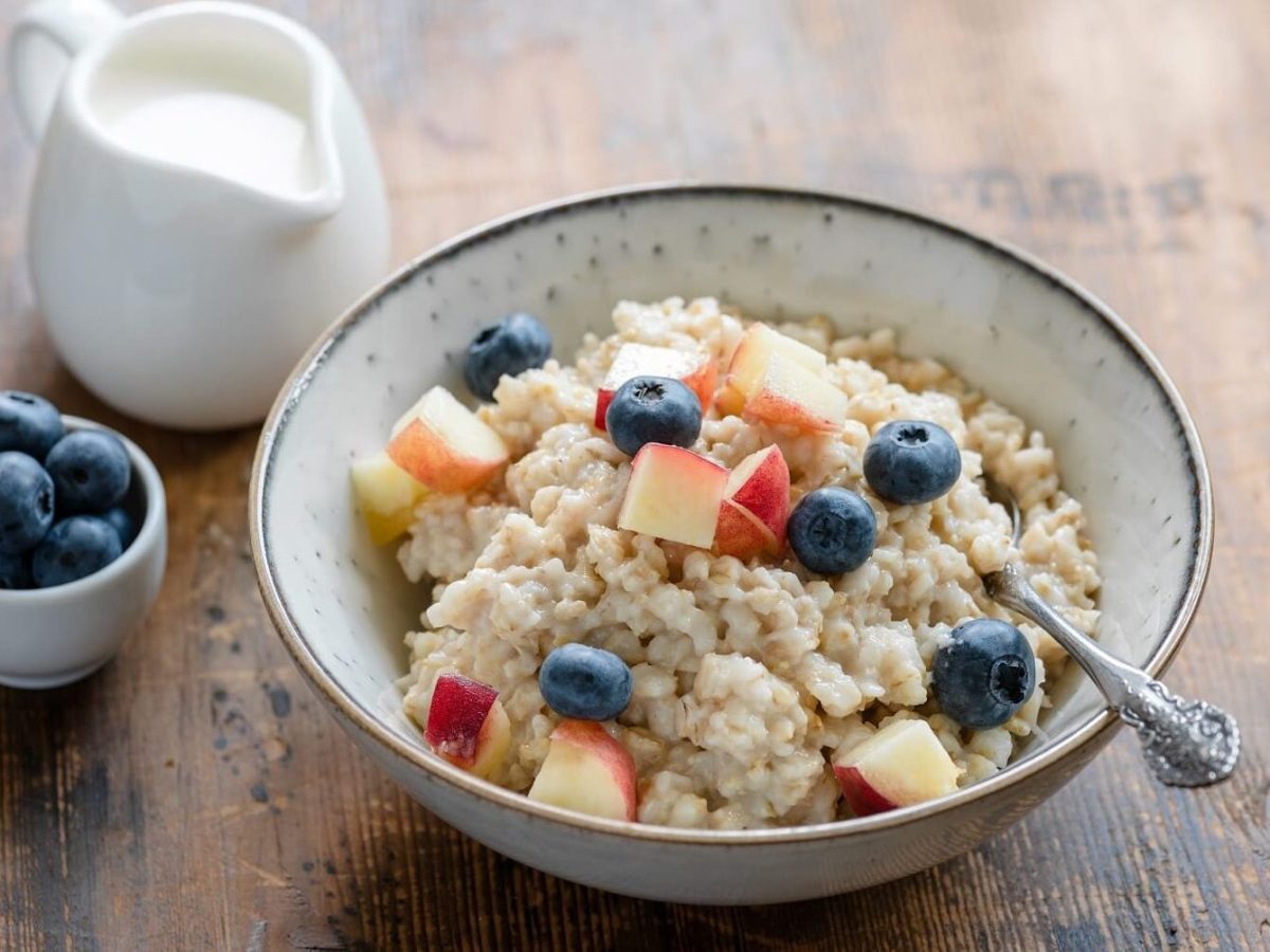 Veganes Bircher Müsli mit Blaubeeren und Pfirsich in einer hellen Schüssel auf einem Holztisch neben einem Kännchen mit Milch und einer kleinen Schüssel mit Blaubeeren.