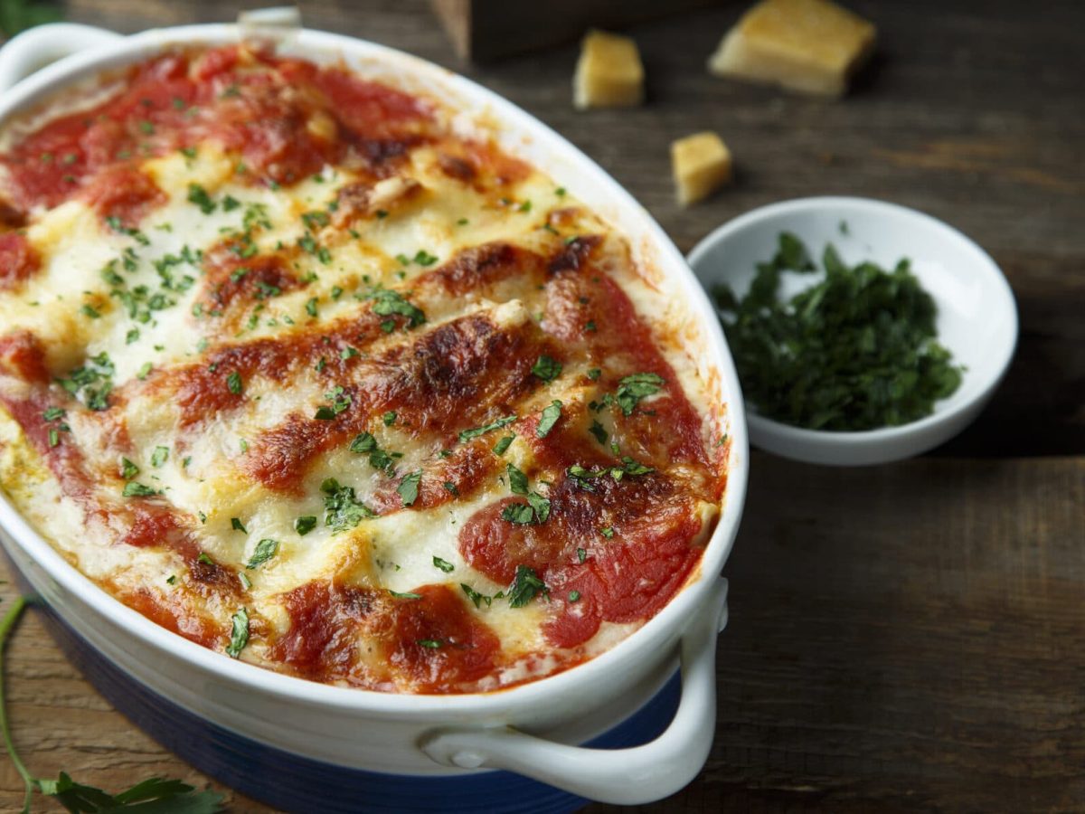 Vegetarische Cannelloni überbacken in einer Auflaufform, daneben eine Schale mit Kräutern und ein paar Stücke Parmesan.