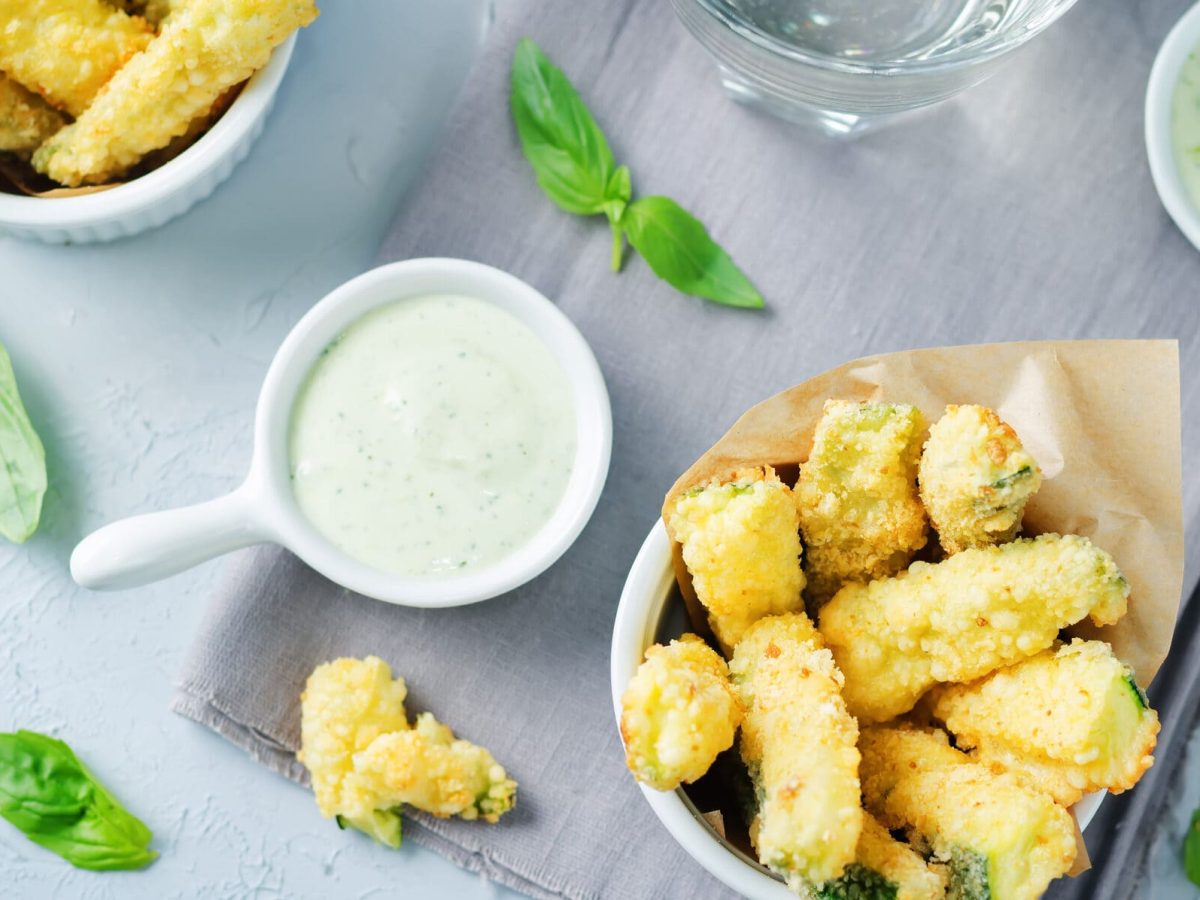 Hellblauer Untergrund mit einer Schale Zucchini-Pommes umgeben von Dips.