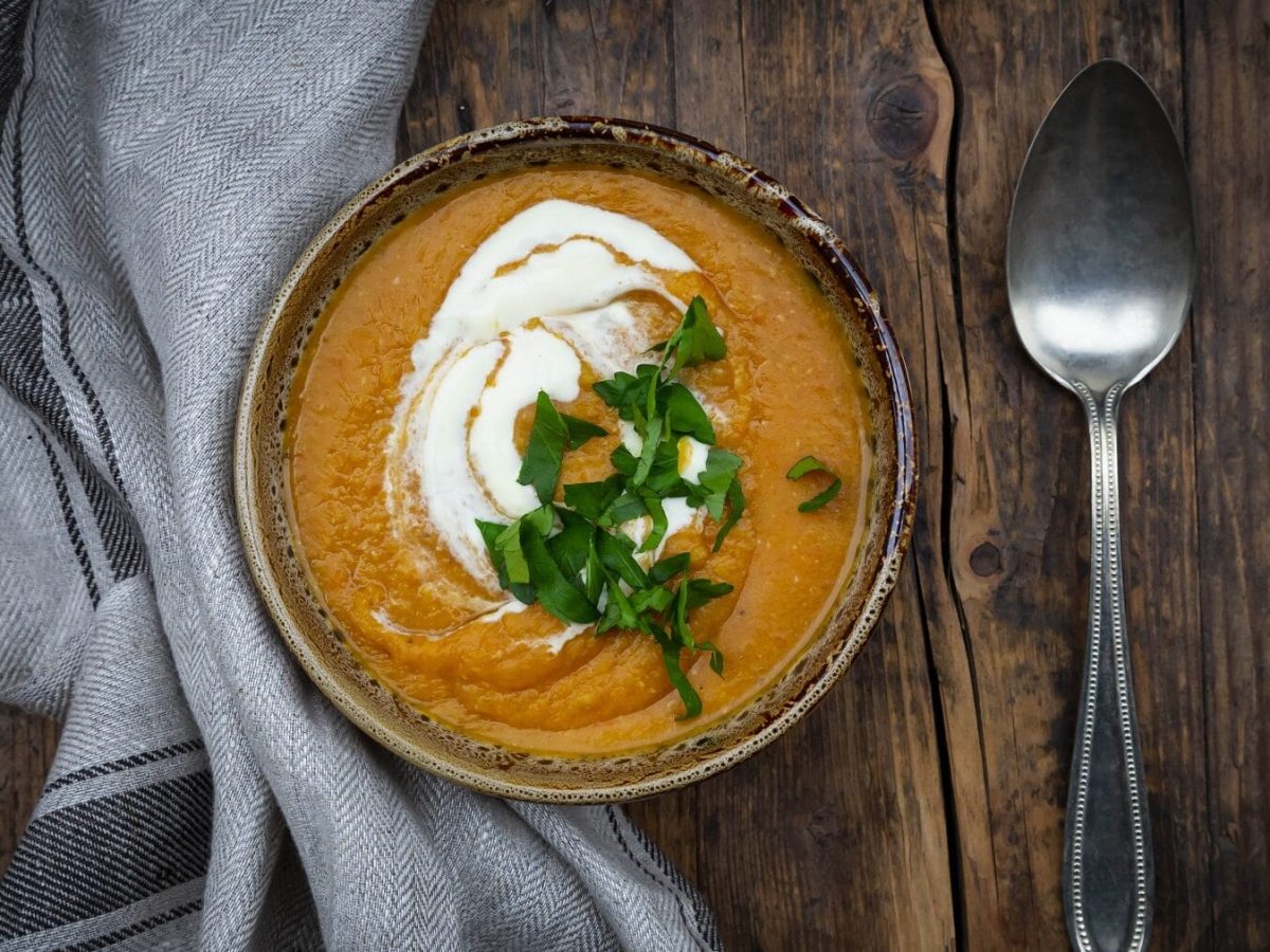 Rote Linsensuppe mit Créme Fraiche und frischer Petersilie in einer gemusterten Schüssel auf einem dunklen Holztisch neben einem grauen Tuch un einem Löffel.