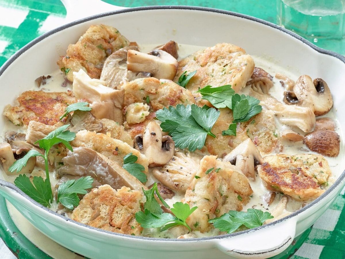 Eine weiße Pfanne mit veganem Semmelknödel-Geschnetzelten mit Pilzen steht auf einem weiß-grünen Untergrund. Rechts oben neben der Pfanne steht ein Glas mit Wasser. Das Geschnetzelte ist garniert mit Petersilie.