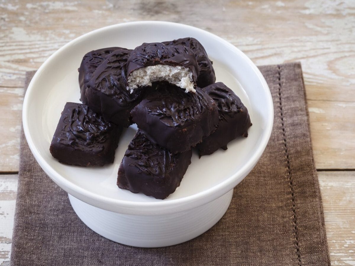 Einige selbst gemachte vegane Schokoriegel mit Kokos liegen gestapelt auf einem weißen Kuchenpodest. Das Podest steht auf einem braunen Handtuch auf einem hölzernen Untergrund. Der oberste Schokoriegel ist angebissen, sodass seine helle Füllung sichtbar ist.