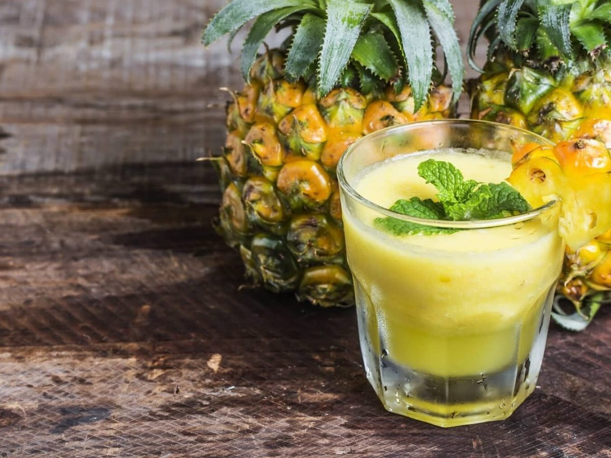 Ein Ananas-Smoothie in einem klekinen Glas steht auf einem Holztisch. Hinter dem Glas stehen zwei Ananasfrüchte. Der Smoothie ist angerichtet mit einer Scheibe Ananas und etwas Minze.