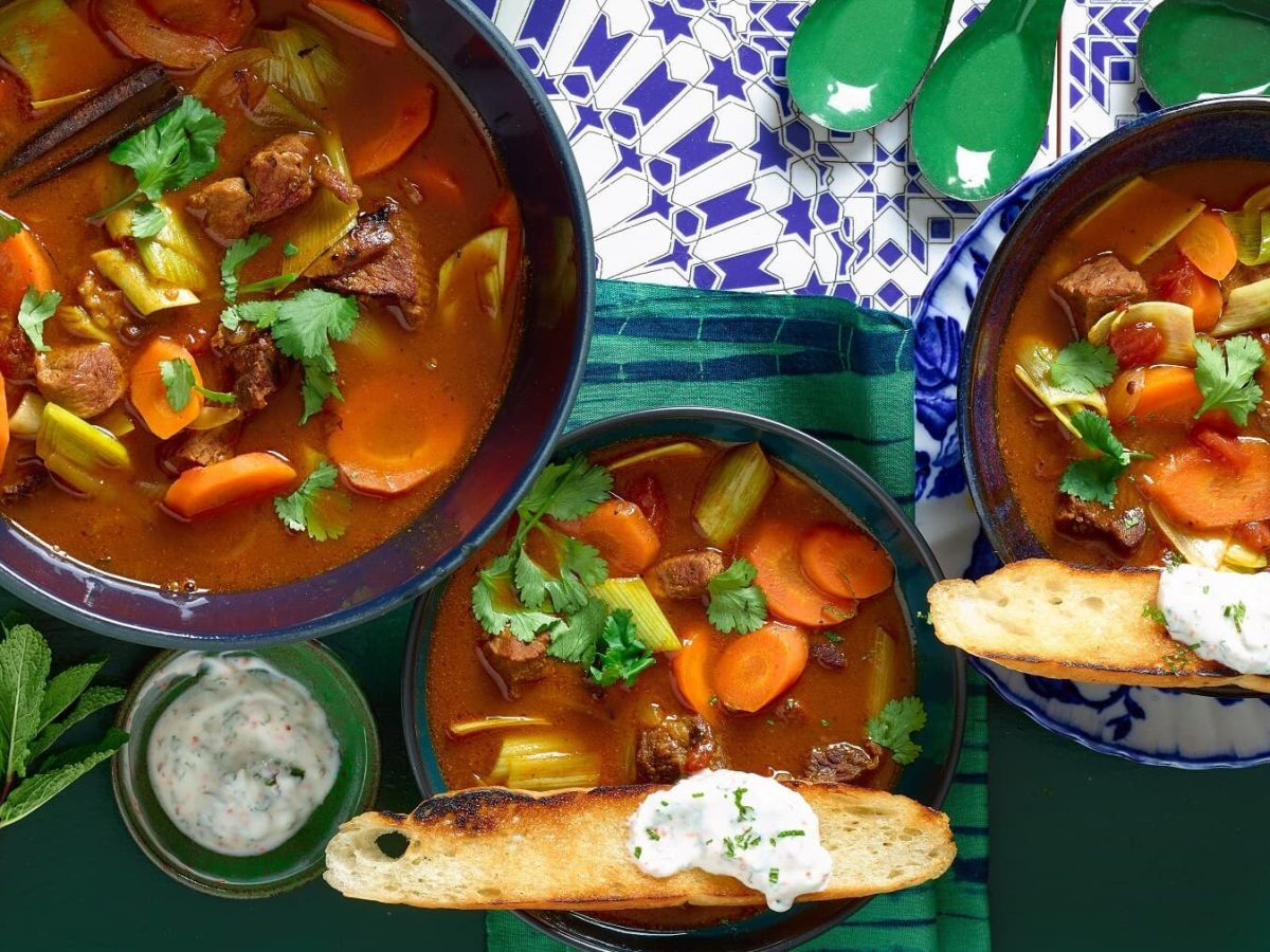 Arabische Gulaschsuppe in drei verschiedenen Tellern mit Brot und Dip auf einem Fliesentisch mit Löffeln und einem Brotkorb.