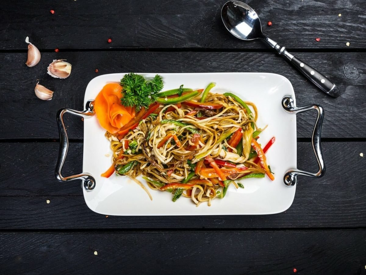 Eine Portion asiatischer Bratnudeln liegt auf einem weißen Tablett mit silbernen Griffen auf einem schwarzen Holztisch. Neben dem Tablett liegt ein Löffel und drei Knoblauchzehen. Auf dem Tisch sind Pfefferkörner verteilt.