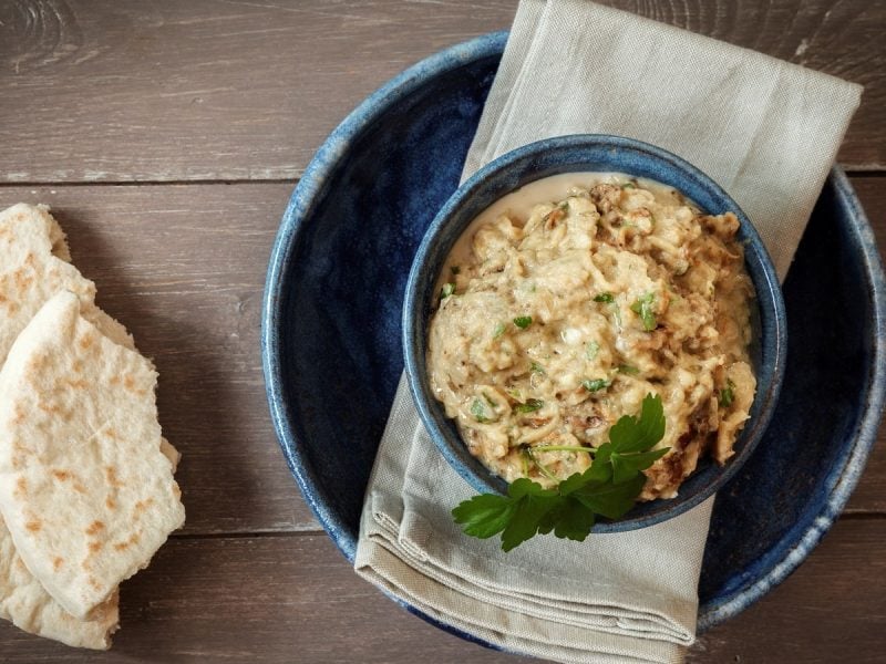 Eine blaue Schüssel mit Baba Ghanoush gefüllt und Petersilie verziert neben zwei angebrochenen Stücken Fladenbrot, von Oben vor einem Holzhintergrund.