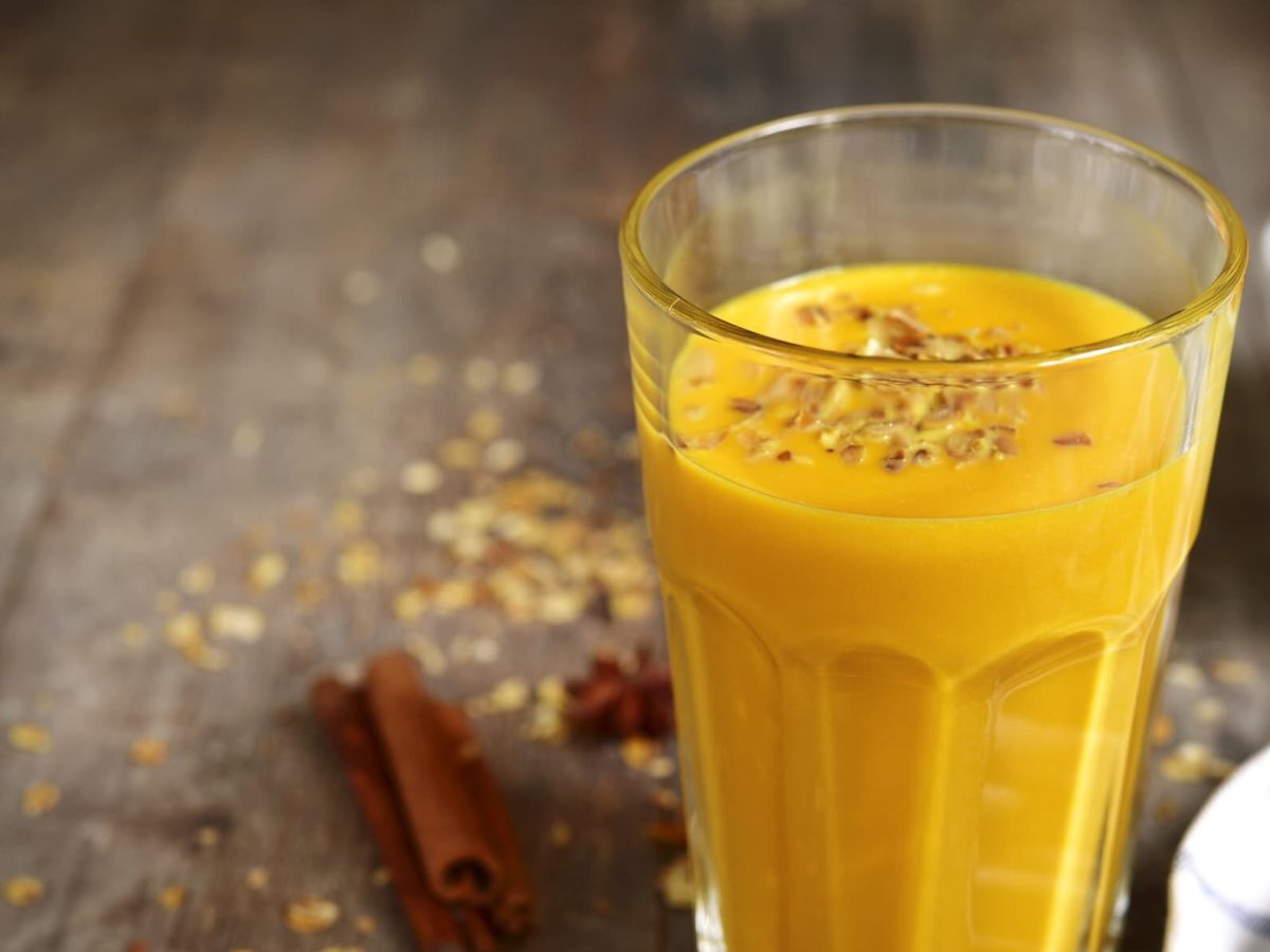 Bananen-Kürbis-Smoothie im Glas auf einem Holztisch, daneben Zimtstangen und Sternanis, ein Geschirrtuch und eine Schale mit Kürbisstücken.