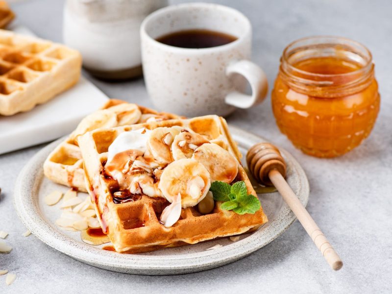 Bananewaffeln mit Bananenscheiben, Mandeln, Joghurt und Minze auf einem grauen Teller neben einem Honiglas, einer Kaffeetasse und einem Brett mit Waffeln auf einem grauen Steintisch.