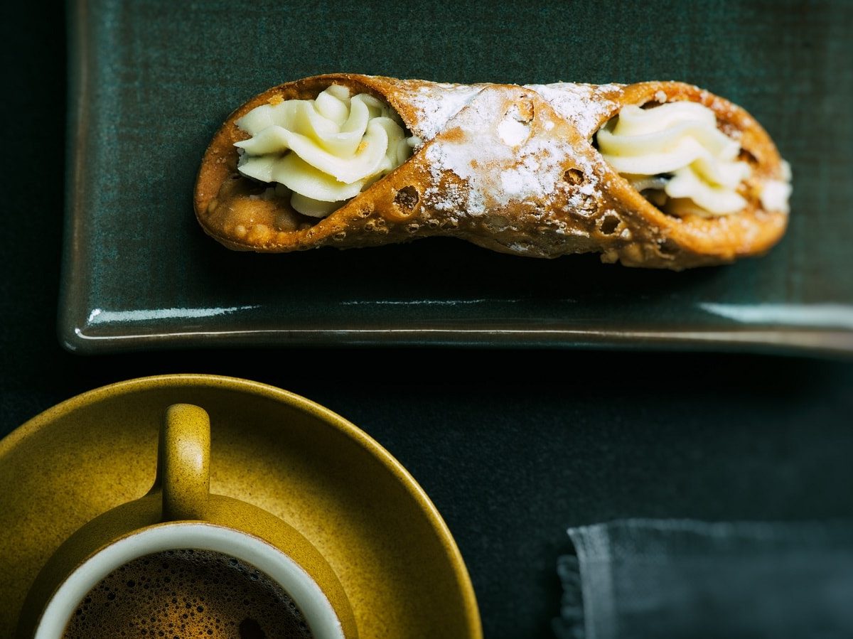 Ein sizilianisches Cannoli mit Puderzucker bestäubt auf einem rechteckigen, dunklen Teller neben einer Tasse Espresso vor dunklem Hintergrund von oben.