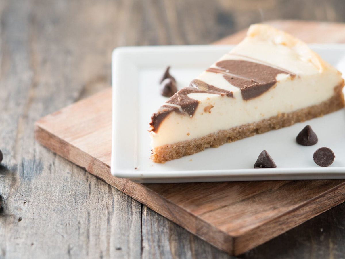 Ein Stück Cheesecake mit Schoko-Swirl auf einem Desserteller, der auf einem Holzbrett steht. Daneben ein paar Schoko-Drops