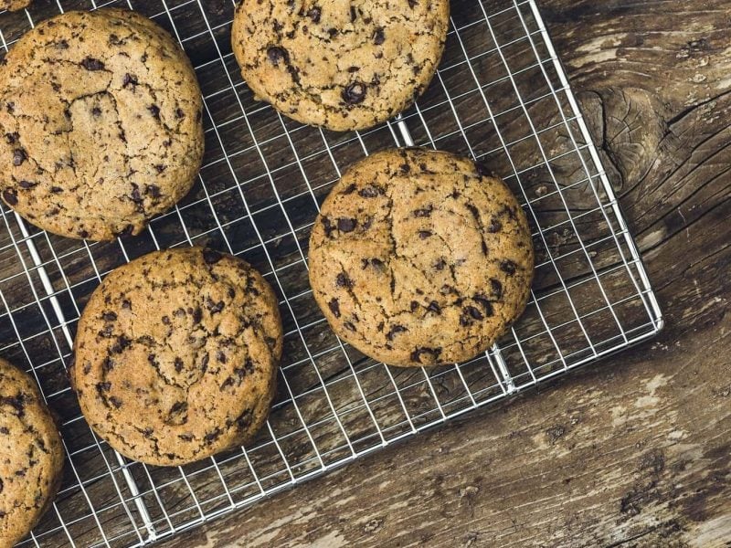 Mehrere runde Chocolate-Chip-Cookies liegen auf einem silbernen Gitter auf einem braunen Holztisch.