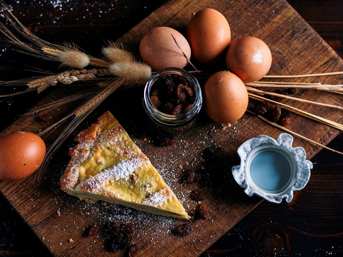 Ein Stück der Crostata Ricotta e Visciole mit Puderzucker bestreut, neben Eiern, einem Glas mit Kirschen und getrockneten Weizenstängeln, auf einem Holzbrett von Oben.