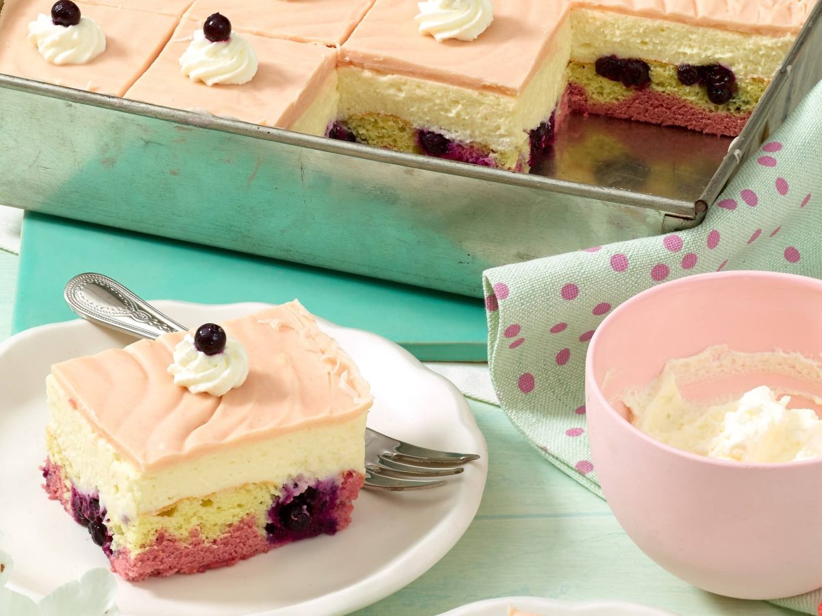 Donauwelle mit Marzipan aus einem tiefen Blech auf einem weißen Teller neben einer rosa Schüssel mit Sahne auf einer grünen Tischdecke.