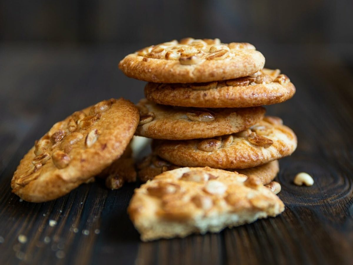 Erdnuss Cookies liegen gestapelt auf einem braunen Holztisch. Fünf Kekse bilden einen Turm, ein sechster Keks ist seitlich gegen den Turm gelehnt. Im Vordergrund liegt ein halbierter Keks und einige Krümel.