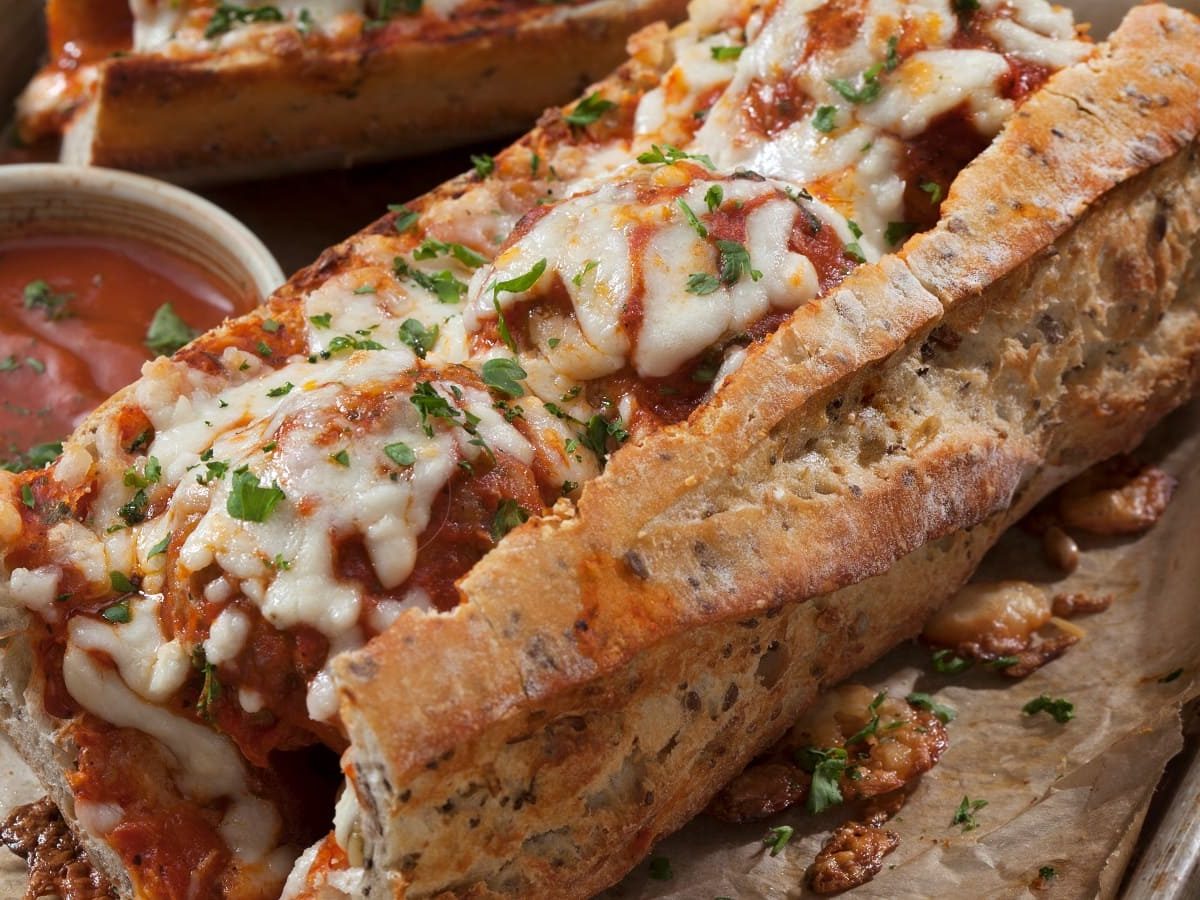 Zwei überbackene Baguette-Sandwiches mit Fleischbällchen und Tomatensauce, garniert mit Basilikum neben einer kleinen Schüssel Tomatensoße.