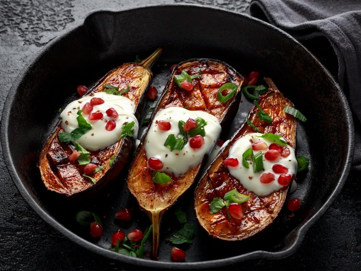 Gebackene Augerginen in einer tiefen schwarzen Pfanne mit Joghurt, Granatapfel und Lauchzwiebeln auf einem schwarzen Tisch neben einem schwarzen Tischtuch.