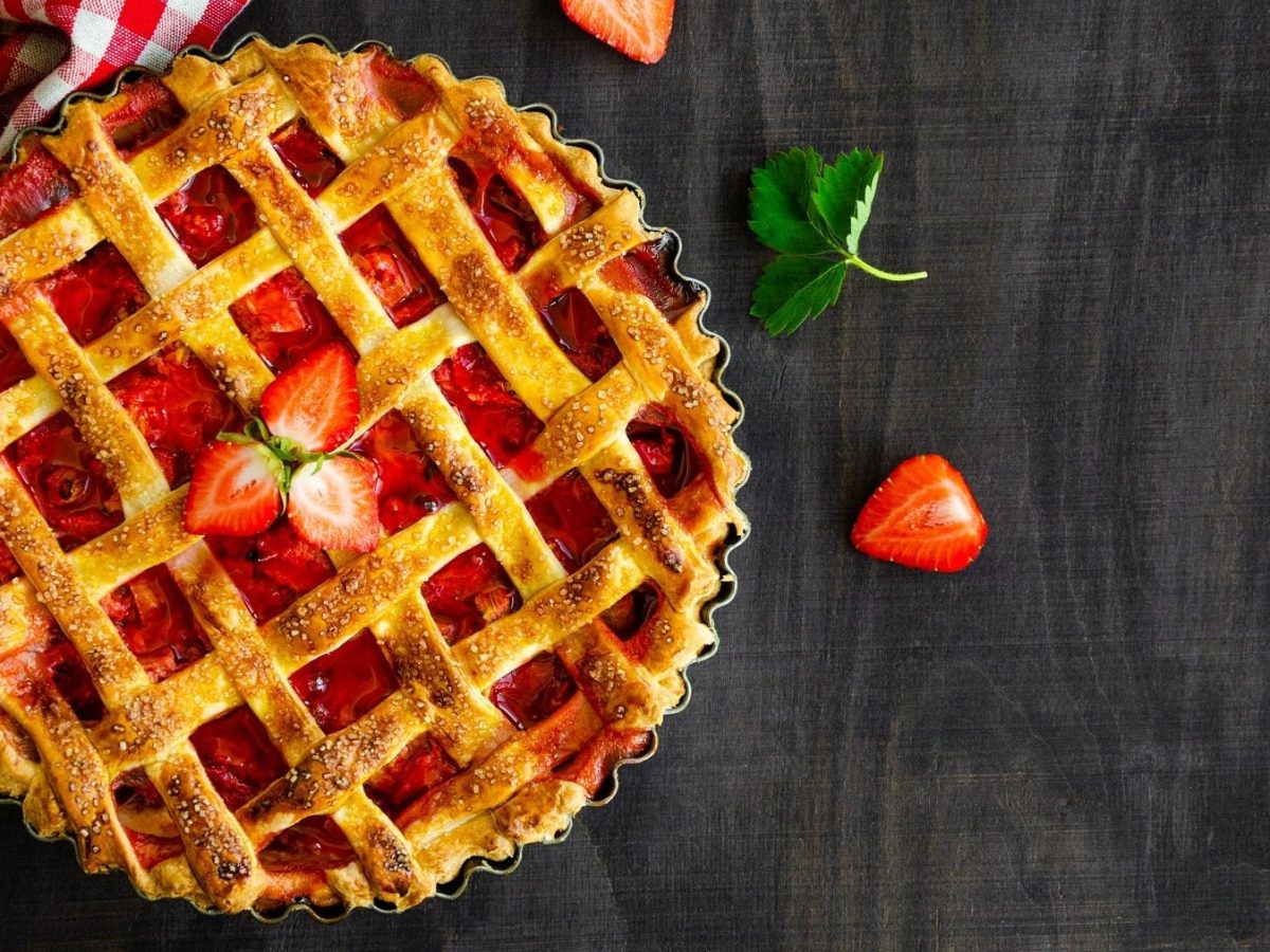 Gitterkuchen mit Erdbeeren und Rhabarber in einer Kuchenform auf einem schwarzen Holztisch neben einer karierten Tischdecke.