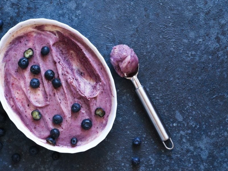 Eine weiße Schale mit Heidelbeer-Buttermilch-Sorbet steht auf einem blauen Untergrund. Das Sorbet ist angerichtet mit einigen Heidelbeeren. Neben der Schüssel liegt ein Eisportionierer mit einer Portion Sorbet.