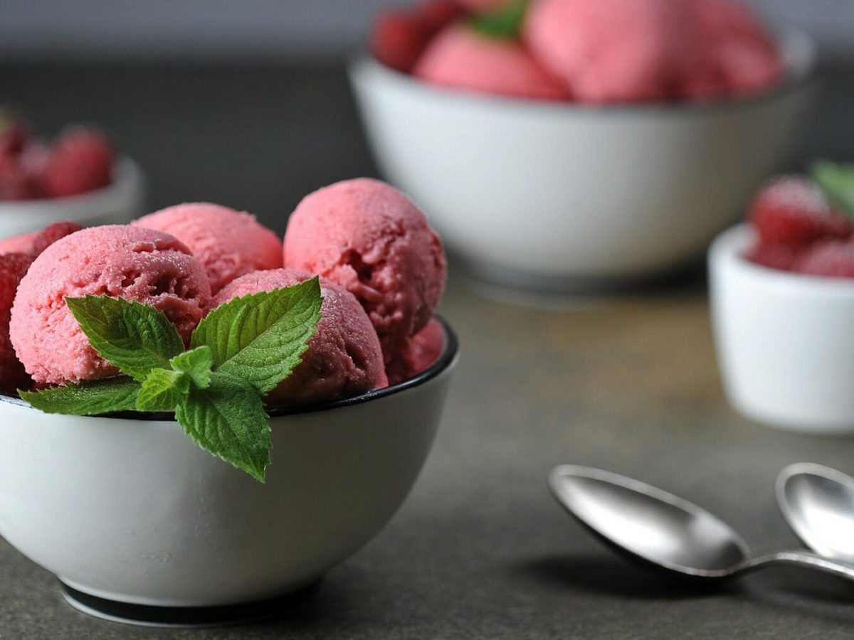 Mehrere Schüsseln mit Himbeer-Joghurt-Sorbet stehen auf einem grauen Untergrund. Die Schüsseln sind weiß und das Sorbet ist mit Minzblättern und Himbeeren garniert. In der rechten unteren Bildecke liegen zwei Löffel.