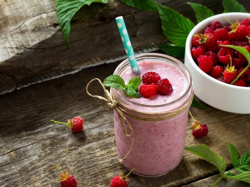 Himbeer-Smoothie mit Banane in einem Glas mit frischer Minze und Himbeeren auf einem Holztisch neben einer Schüssel mit Himbeeren.