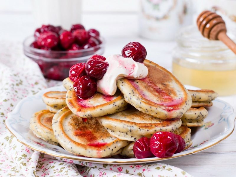 Kirschpfannkuchen mit Mohn und einem Klecks sind serviert auf einem Teller mit Goldrand. Die Pfannkuchen sind mit etwas Sahne angerichtet. Im Hintergrund liegt eine Schüssel mit Sauerkirschen und eine Schüssel mit Honig.