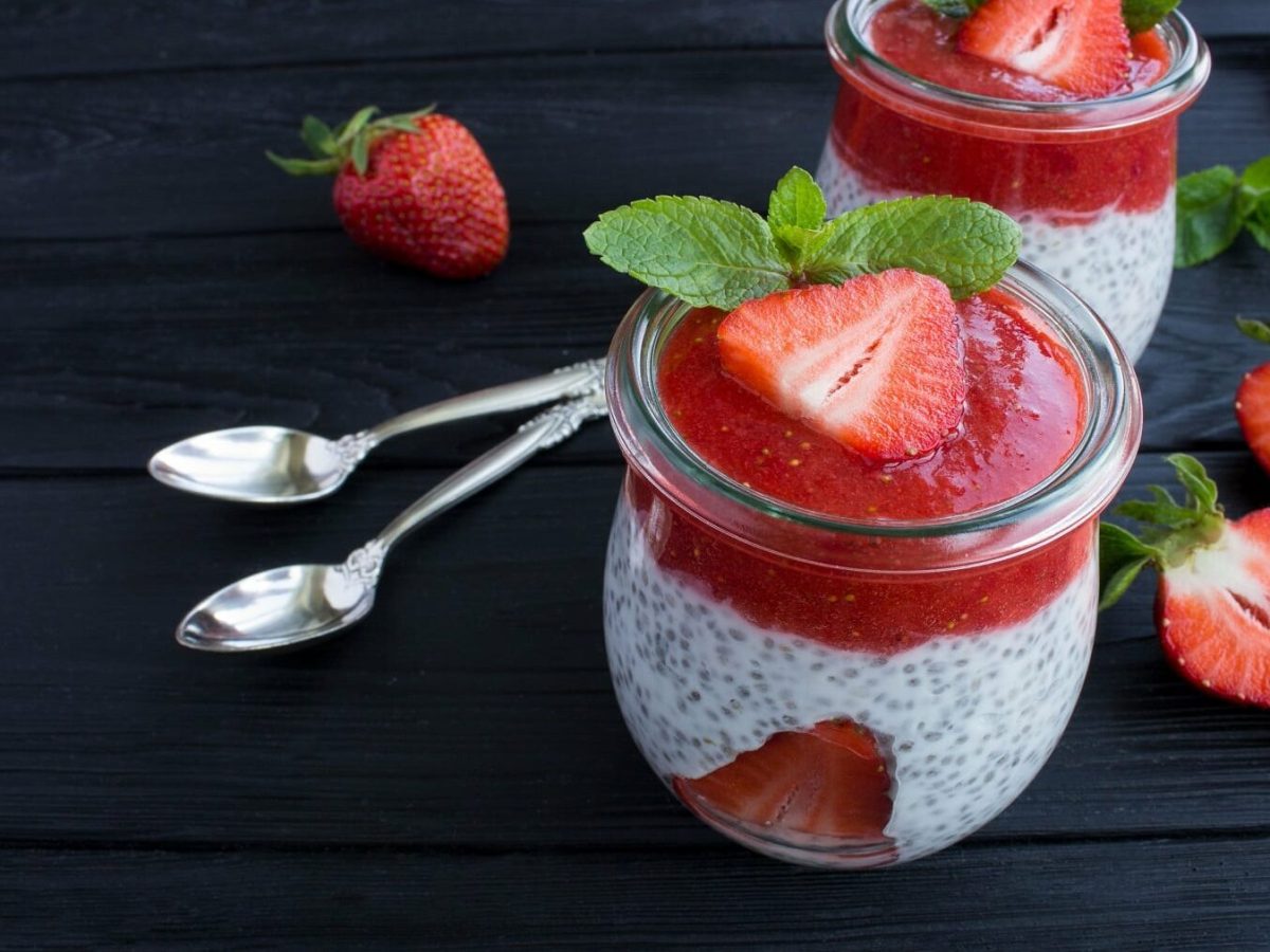 Minzpudding mit Chia und Erdbeeren in kleinen Dessertgläsern neben Löffen auf einem dunklen Tisch.