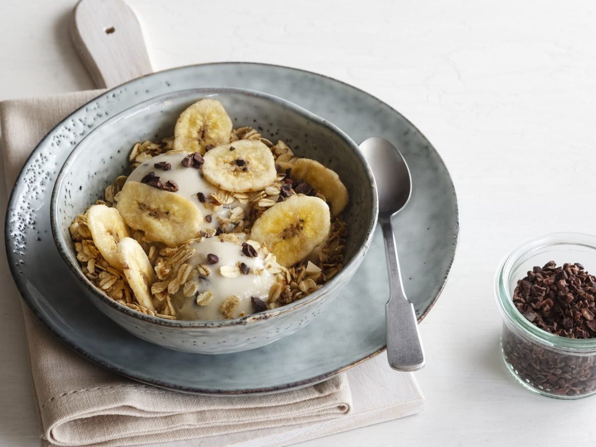 Nicecream mit Bananen und Haferflocken in einer grau-blauen Schüssel mit Löffel auf einem Teller neben einem Gläschen mit Kakaonips auf einem weißen Tisch.