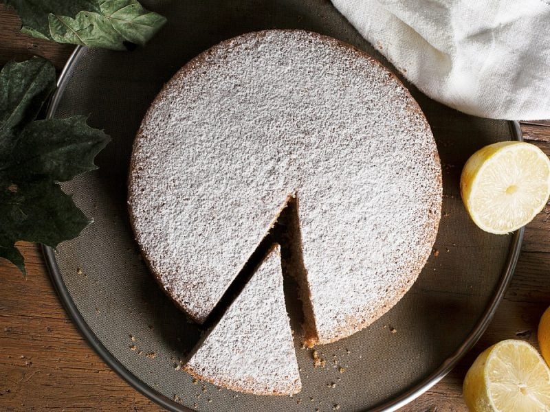 Ein runder mit Puderzucker bestreuter Olivenöl-Zitronenkuchen mit einem Stück eingeschnitten auf einem Backblech, neben halbierten und ganzen Zitronen, Blättern und einem weißen Leinentuch, von oben vor einem Holzhintergrund.