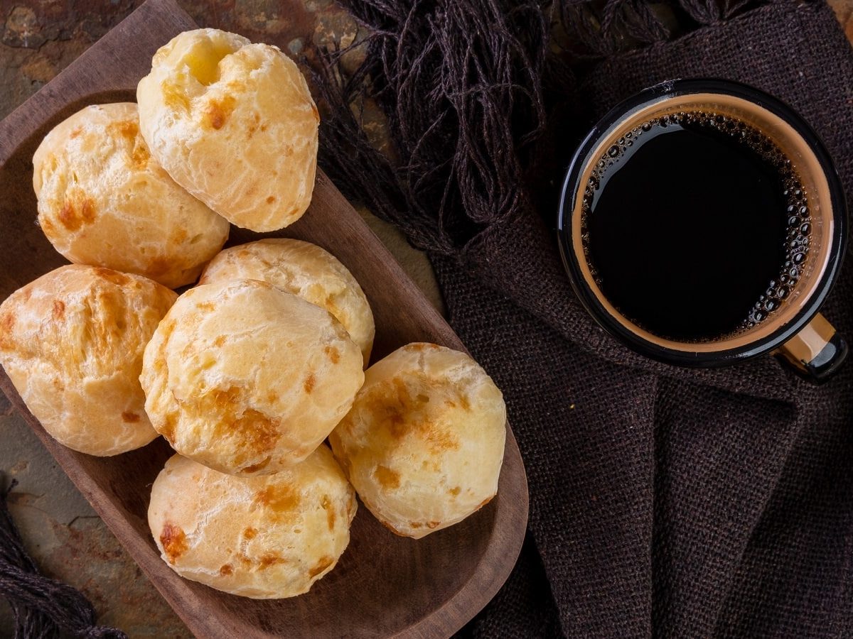 Eine Holzschale mit mehreren Pão de Queijo neben einer Tasse Kafee, von oben, vor dunklem Hintergrund.