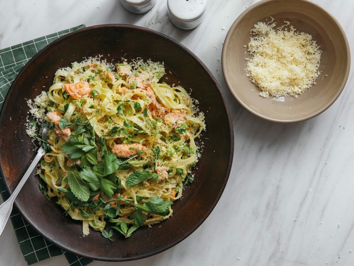 Ein großer, brauner Teller gefüllt mit einer Pasta-Lachs-Pfanne steht auf einem grün karierten Geschirrtuch auf einem Mamortisch. In der Schüssel steckt eine Gabel. Neben der Schüssel steht eine weitere, kleinere Schüssel mit Parmesan und jeweils ein Salz- und Pefferstreuer.