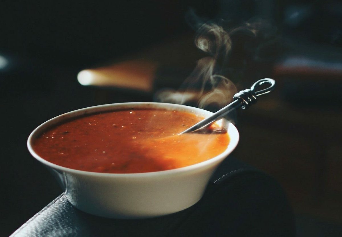 Ein Teller mit dampfender, pinkanter Tomatensuppe steht auf der Lehne eines Stuhls in einem dunklen Büro. In der Suppe liegt ein Löffel.