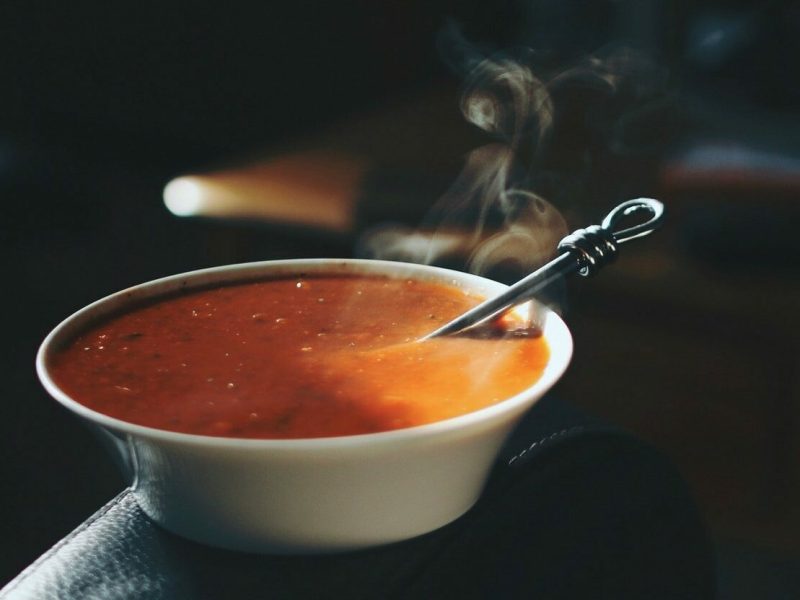 Ein Teller mit dampfender, pinkanter Tomatensuppe steht auf der Lehne eines Stuhls in einem dunklen Büro. In der Suppe liegt ein Löffel.