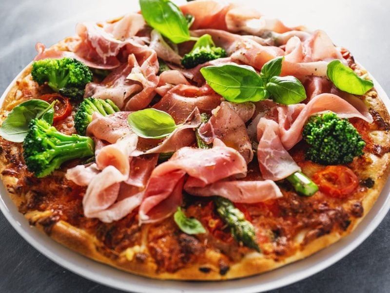 Eine Pizza mit Schinken und Brokkoli steht auf einem weißen Teller auf einem grauen Tisch. Die Pizza ist angerichtet mit frischen Basilikumblättern.