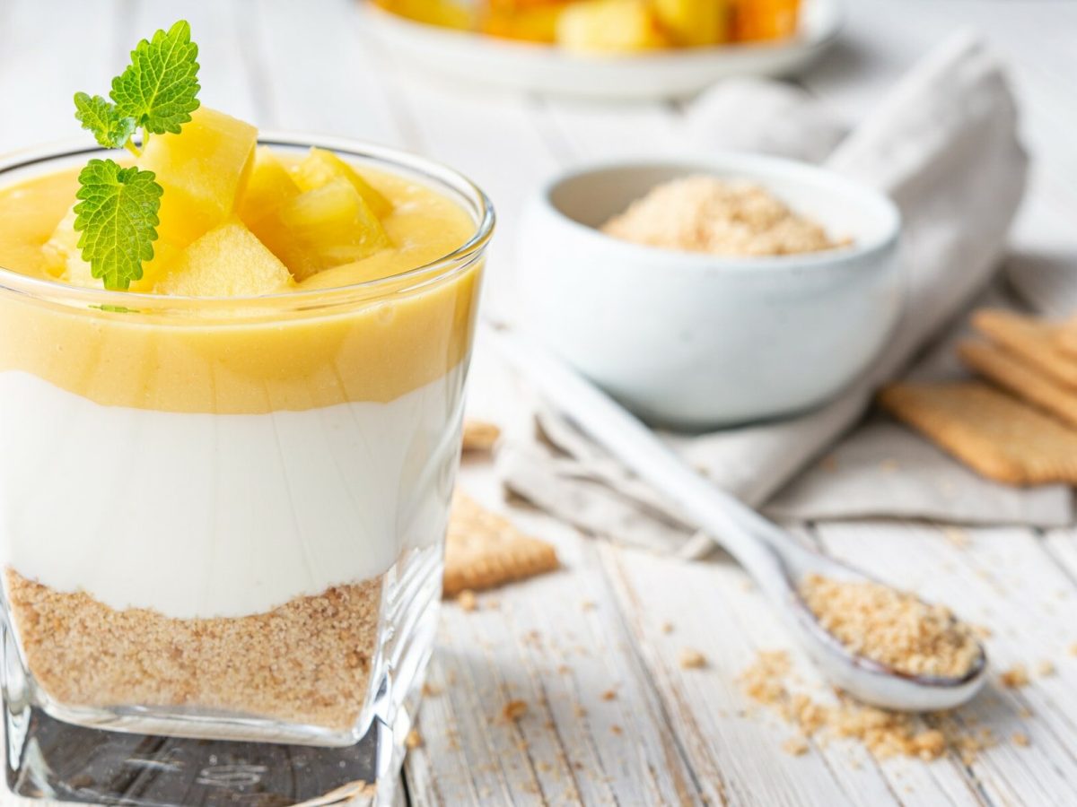 Ricotta-Creme mit Apfelkompott im Dessertglas auf weißem Holz, im Hintergrund Butterkekse, ein Löffel und eine Schale mit Kekskrümeln.