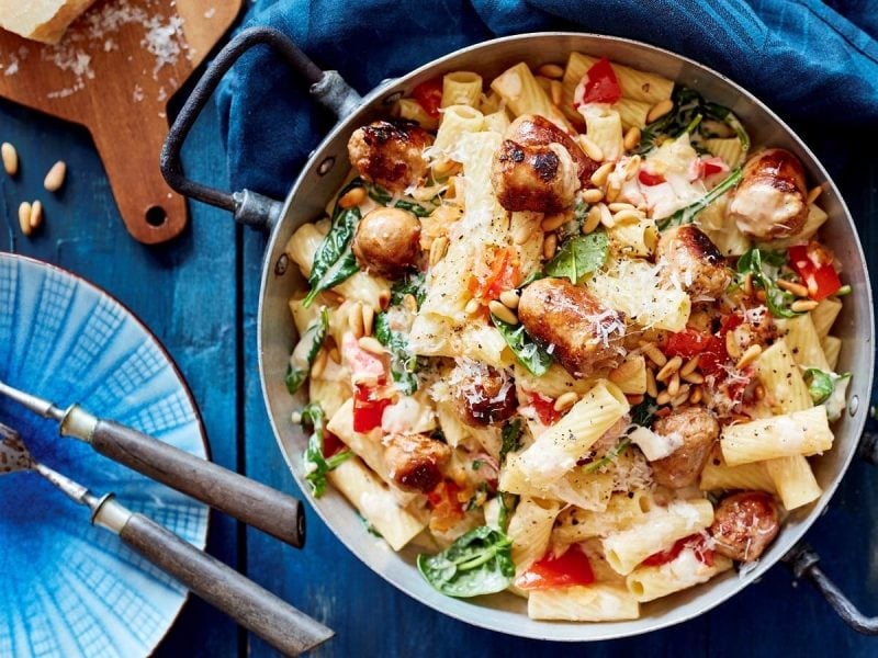 Ein großer Kochtopf mit den Rigatoni mit Salsiccia, Tomaten und Spinat und Pinienkernen neben einem Teller mit Gabel und Parmesankäse, von oben vor blauem Hintergrund.