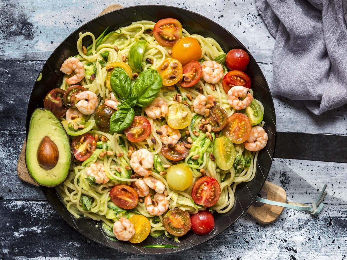 Spaghetti mit Garnelen, Avocado und Tomaten in einer schwarzen Pfanne auf einem gräulichen Holztisch neben einer grauen Tischdecke.