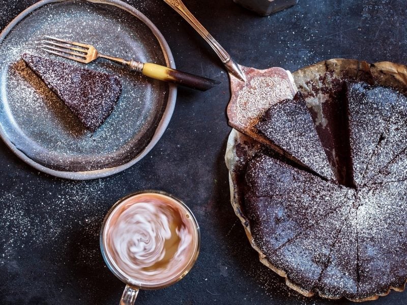 Eine ganze Torta Barozzi mit Puerzucker bestreut und in Stücke geschnitten neben einem Teller mit einem Stück des Kuchens und einer Tasse mit Kaffee, von oben vor dunklem Hintergrund.