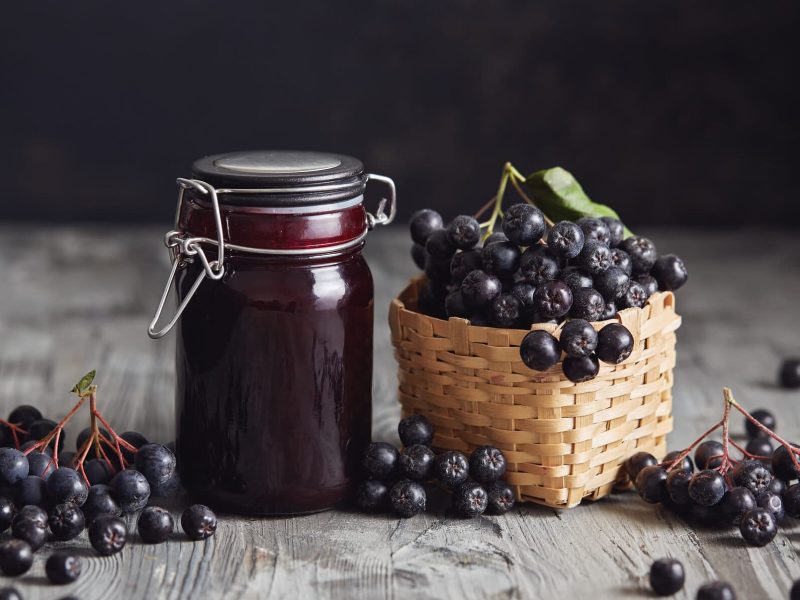 Ein Glas Aroniamarmelade mit Rotwein, daneben steht ein kleines Körbchen mit Aroniabeeren.