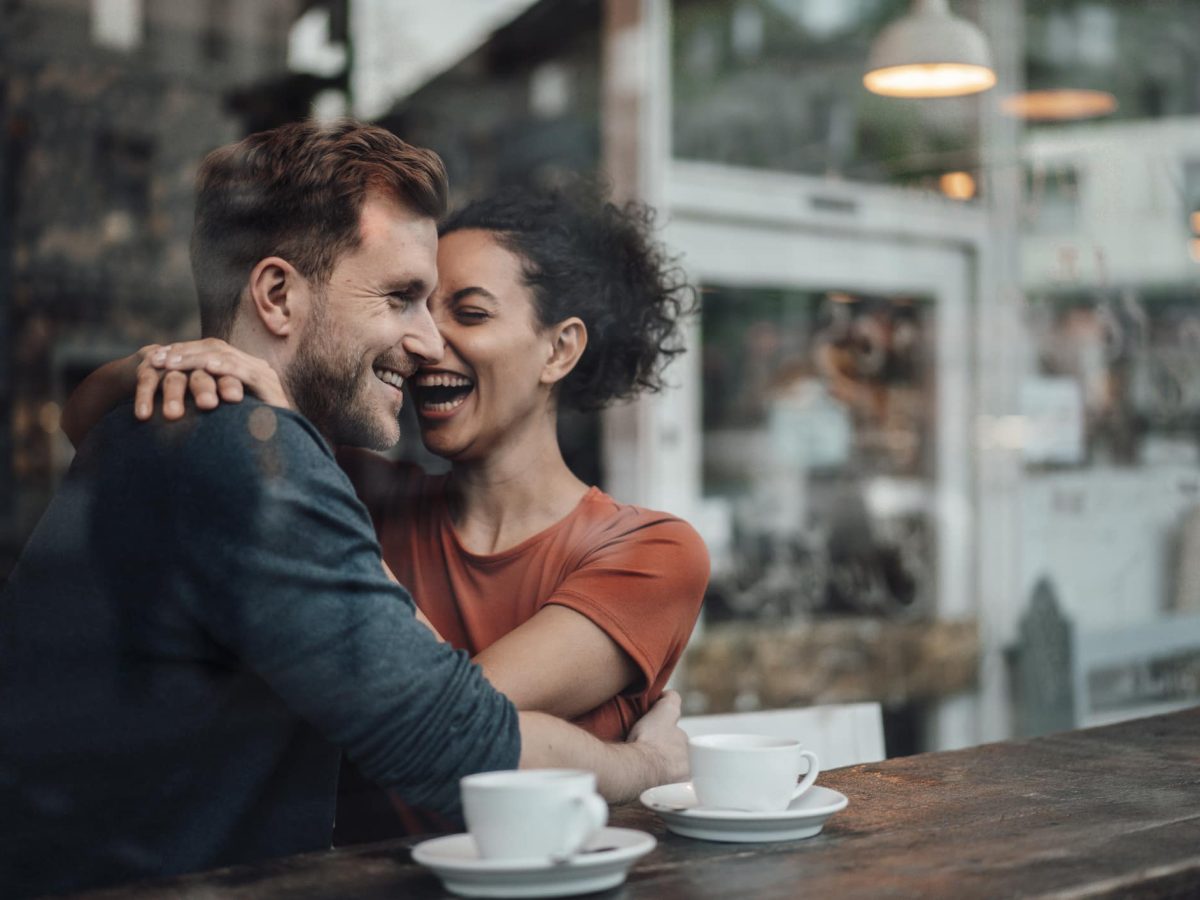 Bumble Brew: Hinter der Glasscheibe eines Cafés umarmt sich ein lachendes Pärchen bei zwei Tassen Kaffee.