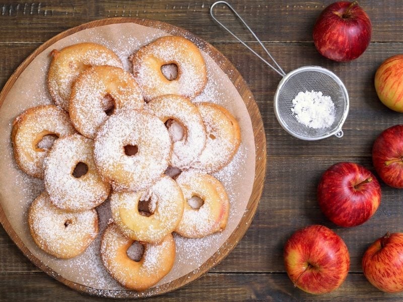 Ein rundes Holzbrett mit aufeinandergestapelten Frittelle di Mele, die mit Puderzucker bestreut sind, neben ganzen Äpfeln und einem kleinen Sieb vor einem Holzhintergrund von oben.