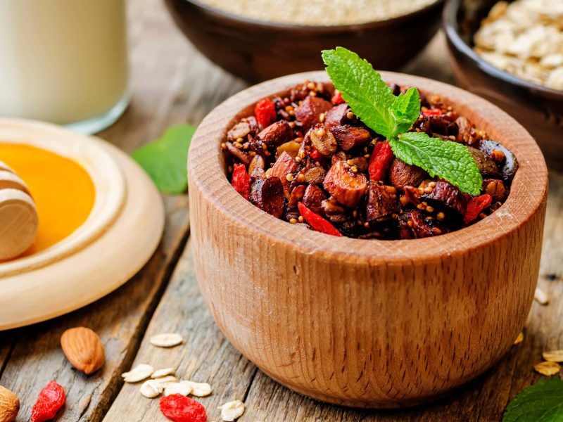 Granola mit Quinoa und Heidelbeeren in einer hellen Holzschüssel neben Honig und Haferflocken auf einem hellen Holztisch.