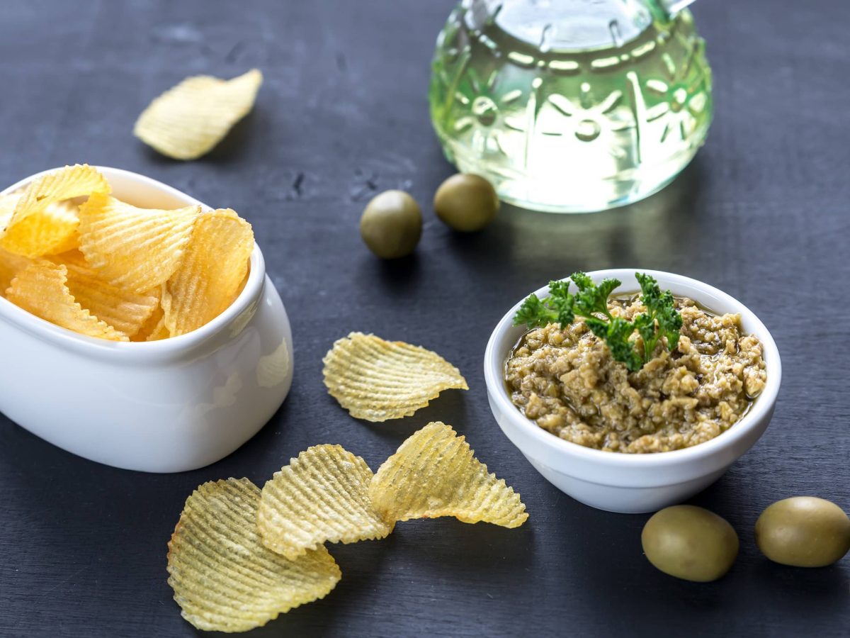 Grüne Oliventapenade in einer kleinen weißen Schüssel auf einem schwarzen Holztisch neben kartoffelchips, frischen Oliven und einer Karaffe mit Weißwein.