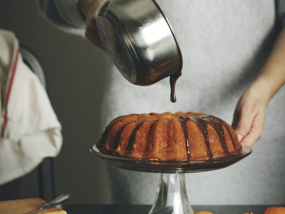 Ein Kuchen steht auf einem Kuchenständer auf einem schwarzen Tisch. Auf dem Tisch liegen Mandarinen und ein Holzbrett. Eine Frau in einem Wollkleid gießt Schokoglasur über den Kuchen.