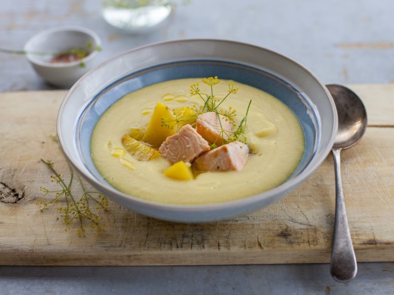 Kartoffelsuppe mit Lachs in einer Schüssel. Daneben ein Löffel. Das Ganze steht auf einem Holzbrett.