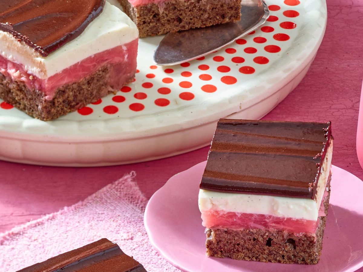 Schokoladige Rhabarberschnitten mit weißer creme auf einem rosa Teller mit einer Gabel auf einem rosa Tisch neben einer Servierplatte mit weiteren Stücken Kuchen.