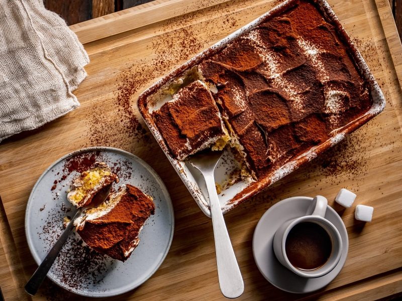 Eine Auflaufform mit klassischem Tiramisu von dem ein Stück herausgeschnitten wurde, das auf dem Teller daneben liegt. Unterhalb der Auflaufform steht eine Tasse Espresso mit zwei Würfeln Zucker. Alles steht auf einem Holzbrett, von oben fotografiert.