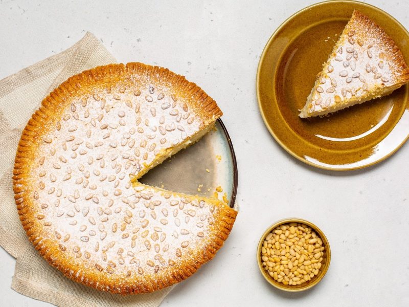 Die runde Torta della Nonna auf einem Teller mit einem Stück abgeschnitten, das auf einem weiteren Teller daneben steht, sowie eine kleine Schüssel mit Pinienkernen, von oben, vor hellem Hintergrund.