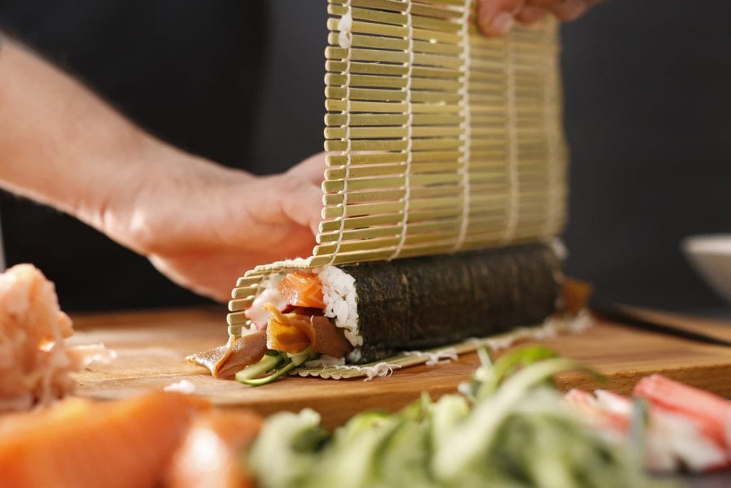 Eine Sushirolle liegt in einer Bambusmatte und wird geformt. Im Vordergrund liegen verschiedene Füllungen für das Sushi